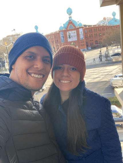 An image of my wife and me, wearing winter clothes from our Airbnb balcony with Campo Pequeno's Praça do Touro in the background, at the day we first got to Portugal